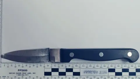 Crown Prosecution Service A black handled kitchen knife, the blade has been snapped away from the hilt but is placed above oit. Below is a ruler measuring the blade and handle to be 195cm long