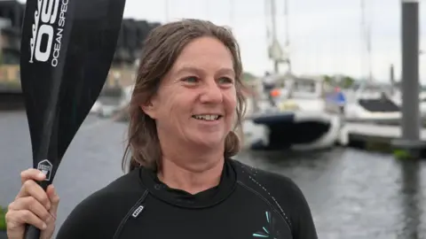 Sarah Perkins holding an ore and wearing a wetsuit. She is stood by some water and is smiling