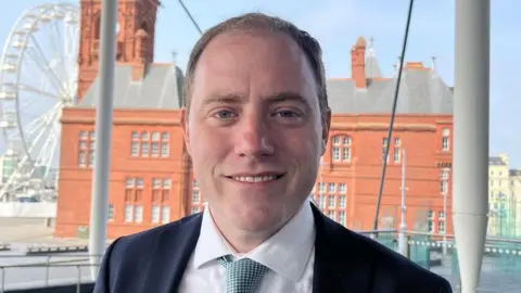 A head and shoulders picture of Rhys ab Owen, wearing a suit and tie and stood on the Senedd steps with a view of Cardiff Bay's Pierhead building in the background.