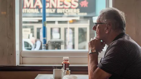 @mwktar Man sitting in a cafe stall with coffee and another shop with a welsh dragon visible through the cafe window