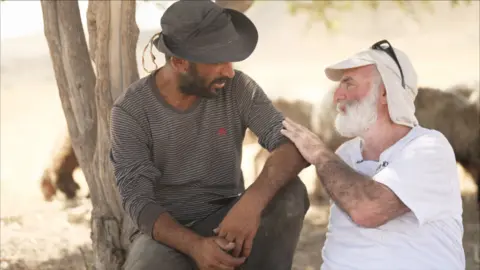 Ahmad, a Palestinian farmer wearing a long sleeved t-shirt and a hat, sitting with Gil, and man with a white beard. Gil has his hand placed on Ahmed's arm and they are in conversation, sitting under a tree.