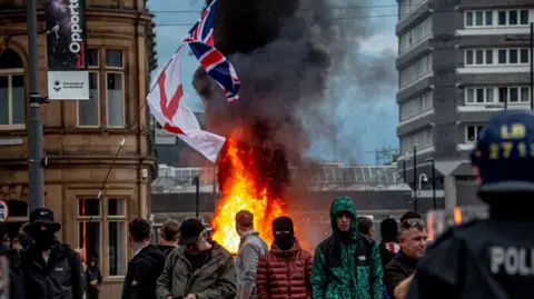 Getty Images A group of men stand in front of a fire during the riots in Sunderland. A union flag and a Cross of St George flies above the scene as police officers look on.