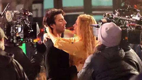 Getty Images Justin Baldoni and Blake Lively pictured on set for It Ends With Us. Justin has floppy brown hair and wears a black jumper. Blake, who has crimped blonde hair, has her arms around his neck looking into his eyes. They're surrounded by camera operators filming them. 