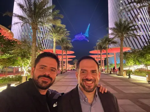 Family Handout Two brothers pose in front of a lit-up decoration shaped like a shark suspended in the sky. They are both wearing jackets and both have black hair with beards.