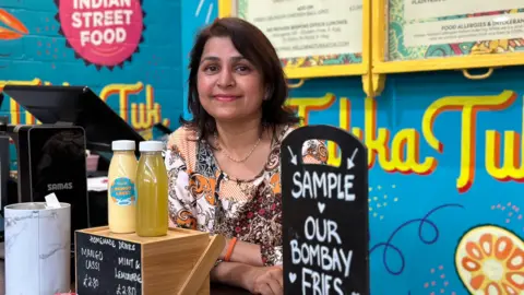 Raphael Sheridan / BBC Rupali smiling at the camera with shoulder length dark hair at the counter of her street food restaurant