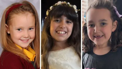 Handout Elsie Dot Stancombe is pictured in school uniform - with maroon cardigan, yellow shirt and grey pinafore dress and matching maroon hairband. Alice da Silva Aguiar is wearing a white lace dress and floral headband, and Bebe King has her hair in bunches and ribbons and is wearing a black T-shirt. All three are smiling at the camera. 