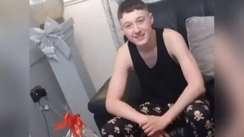 Family photograph A teenage boy smiles at the camera at Christmas. He is wearing pyjama bottoms with gingerbread men on, and a black vest. He has short hair. 