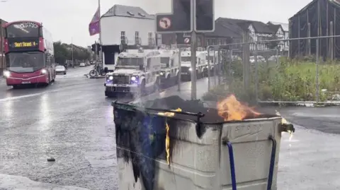 a bin, set on fire, sits on a road in Belfast which is lined with riot vans while a pink bus approaches the area