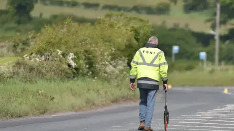 Pacemaker A man wearing a hi-vis jacket with the letters FSNI on the back walking down a road, carring a measuring device