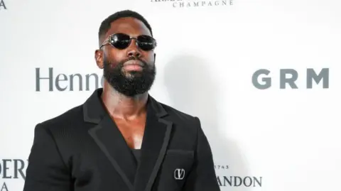 Getty Images Ghetts at the GRM Gala. Ghetts wears a black suit over a black vest top, matched with dark sunglasses. He has a short beard and cropped dark hair. 