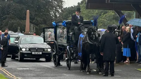 horse-led carriage with coffin arrives in front of people outside chapel