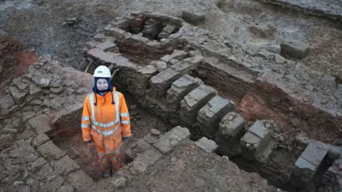 Oxford Archaeology An archaeologist in an orange jump suit with white stripes and a white hard hat standing in the foundations of a Roman tile making unit