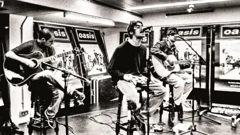 Brian Cannon/Microdot Black and white photo of Bonehead (left), Liam Gallagher (centre) and Noel Gallagher (right) sitting on high stools with microphones in front of them. Bonehead and Noel are playing guitars and Liam is holding a tambourine as he sings into the microphone. There are large Definitely Maybe posters behind them and to the right