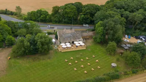 PA Media An aerial shot of a pub, which has picnic tables and umbrellas outside. Next to the pub is a road and there are fields surrounding it.