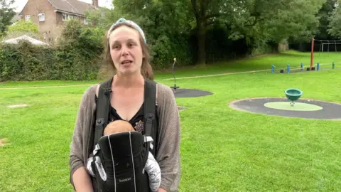 Lauren Slater in a brown cardigan with a baby holder attached with baby standing in front of a park