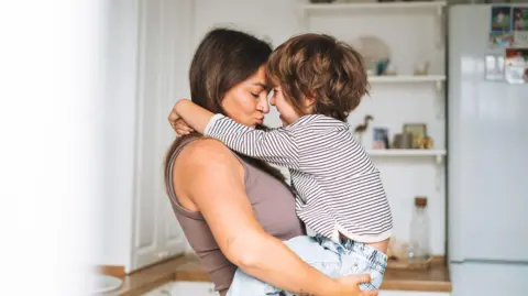 Getty Images Woman carrying young boy affectionately with a fridge and kitchen shelves seen behind