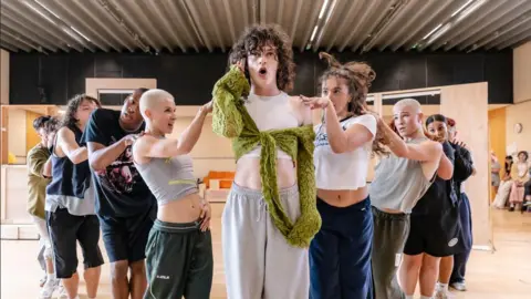 Danny Kaan An actor holds a phone up to their ear while wearing a white tank top and green scarf. They have curly hair. Behind them are six back up dancers in baggy t-shirts and sportswear.