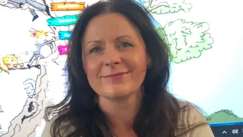 Supplied Anne Marie Kenyon has dark brown wavy hair. She sits in front of a whiteboard in her classroom