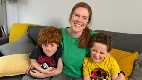 Gemma Laister/BBC Cara Carey sits on a sofa in a green top with her two sons on either side, smiling at the camera 