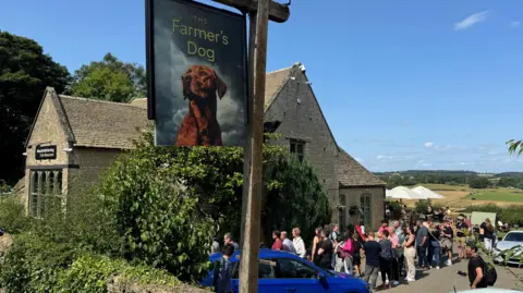 BBC An historic pub under blue skies with rolling fields in the distance and a pub sign with a dog in the foreground