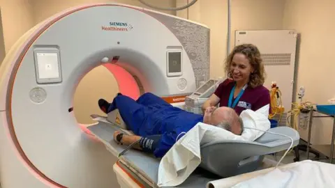 BBC Man lying down about to enter a CT scanner with a nurse next to him