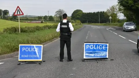 Pacemaker Police officer facing away from camera standing in the middle of the road with two signs that read "Police Road Closed"