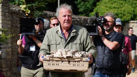 PA Media Jeremy Clarkson, wearing a green coat and holding a tray of mushrooms. There is a camera crew walking behind him