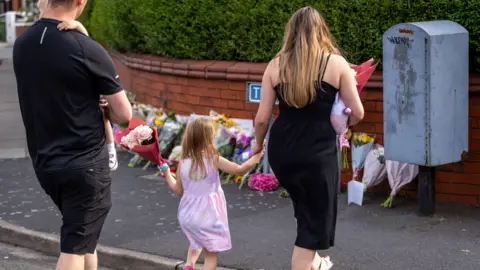 PA Media A young girl in a pink and purple dress lays flowers with her parents