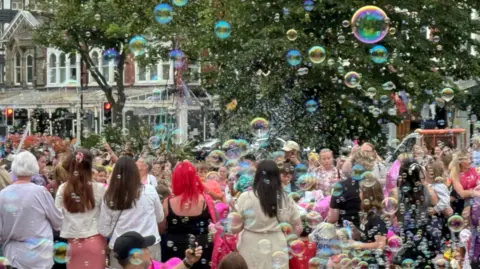 PA Media Children blew bubbles as others placed flowers and heart-shaped balloons in front of The Atkinson arts centre in remembrance of the victims of the stabbing attack