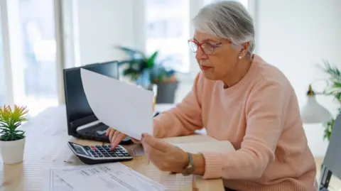 Getty Images Older woman sits at a desk at home holding a bill with her other hand on a calculator and a laptop in the background.