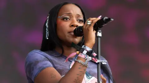 Getty Images Rachel Chinouriri performs during day four of Glastonbury Festival 2024. Rachel is a woman in her 20s with straight black hair. She has brown eyes, made up with gems, and sings into a mic. She wears a purple top and festival wrist bands.