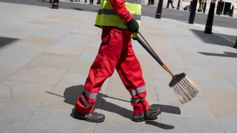 Getty Images Generic image of a street cleaner in London, wearing a red and white uniform and carrying a broom