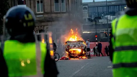 Getty Images Police officers wearing riot gear in Sunderland, viewed from behind close to the camera, as in the middle distance an overturned car burns and a crowd mills around