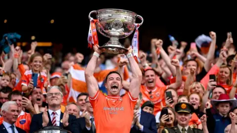 Getty Images A man holding a trophy above his head, he is wearing an orange GAA top with crowds of people cheering behind him