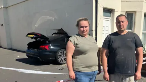 Leanne Rinne/BBC A woman and man stand side-by-side outside of a house with a car buried in the wall