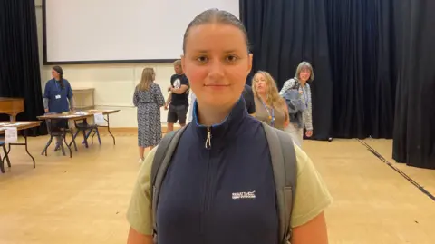 BBC Kingsbridge Community College pupil Anna Petrenko looking at the camera in a sports hall after picking up her A-level results. She is wearing a navy blue gilet jacket and a light green t-shirt and has a back pack on