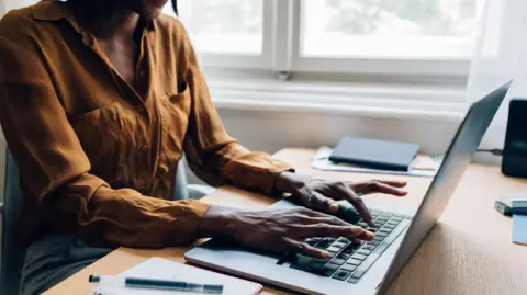 Getty Images An anonymous woman sits at a desk and types on a laptop keyboard