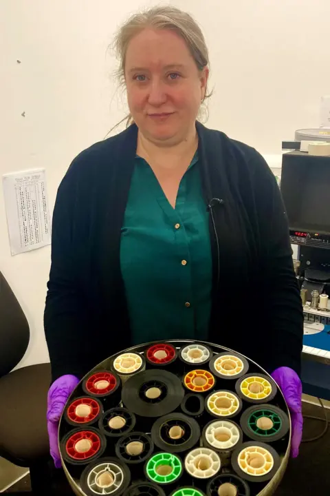 Kay Foubister with some of the film canisters found in the basement at DC Thomson’s Kingsway office. She is wearing purple safety gloves as she handles a large round container with canisters of different colours inside.