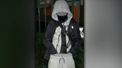 West Midlands Police A boy in a grey tracksuit, with a machete in a black covering tucked into his trousers. His face is blurred and he is standing outside in the dark in front of a fence