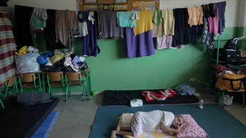 An elderly woman lies on a mattress while clothes hang around her