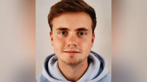  Avon and Somerset Police Photo of Jack who has brown hair, stubble, and is wearing a light blue hoodie.