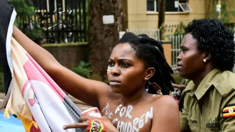 Abubaker Lubowa/ Reuters A Ugandan protester with the words "for the children" painted on her bare chest and holding a flag is detained by a police officer for protesting over corruption outside parliament in Kampala, Uganda - Monday 2 September 2024