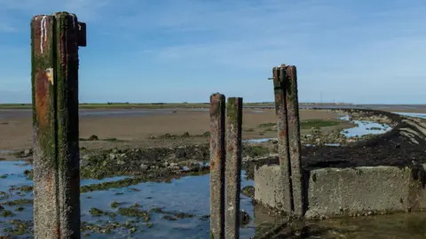 Getty Images Foulness Island. In the foreground there are rusted pillars, which are the remains of an old oil tank, and sea water. In the background there is shingle and clusters of seaweed and rocks