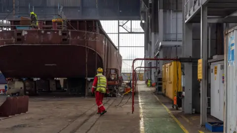 Getty Images The production facility in Harland & Wolff Group Holdings shipyard in Belfast. A man is wearing a yellow hardhat, a high-vis jacket and a red jumpsuit.