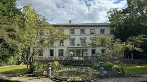 Frannie Brown The front of Fyfield Manor. It is a large white building with pastel green trim. It has a pond complete with lily pads in front of it.