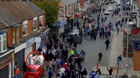 Cleveland Police A mob of people standing in the road. Megan Davison can be seen circled in red standing on top of a red car in the bottom-left corner of the picture.