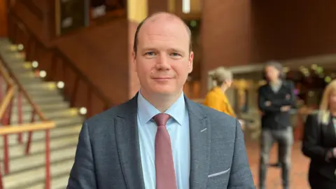 Gordon Lyons wearing a grey blazer, blue shirt and red tie