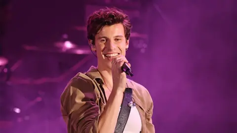 Getty Images Shawn Mendes performing on stage, with a black microphone in his hand, a brown top with a white vest, and a purple haze background.