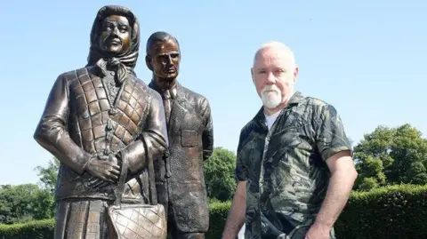 Antrim and Newtownabbey Borough Council Press Release  Anto Brennan posing with his sculpture. He is wearing a khaki sleeveless shirt and beige chinos. He is grey with short hair and a grey beard. 