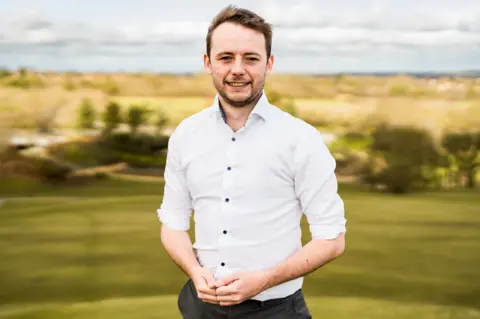 Chris Curtis/MK Labour Labour MP for MK North, Chris Curtis, standing in green space outside in a white shirt. He has short brown hair and stubble and is smiling at the camera.